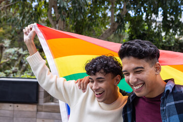 Two men standing on patio holding rainbow pride flag under leafy canopy and planter boxes