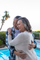 Fototapeta premium Asian couple hugging at edge of turquoise pool showing wristwatch by plant and metal railing