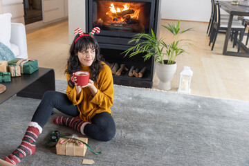 Naklejka premium Woman wrapping christmas gifts on gray rug in living room next to burning fireplace with red mug