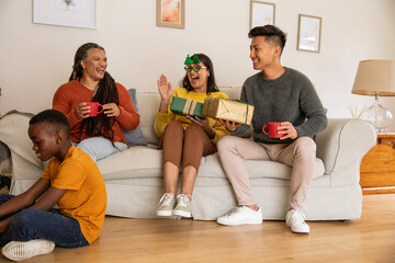 Obraz premium Diverse family sitting at home on couch laughing and holding mugs and gifts, boy playing