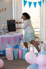 Obraz premium Pregnant african american woman arranging pink, blue cupcakes next to chalkboard in living room