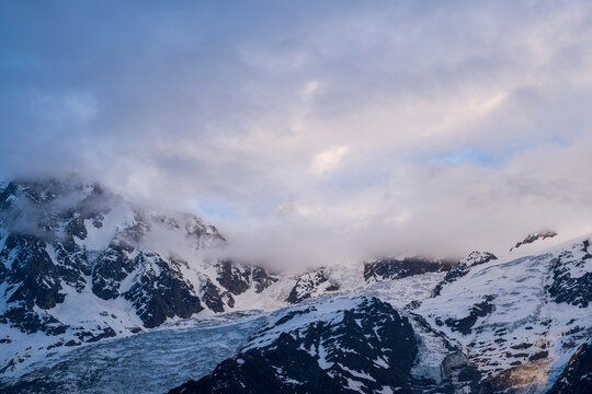 Moody evening clouds drift above the rugged, snow-covered peaks and glacial slopes of Mont Blanc du Tacul, with soft pastel light illuminating the dramatic alpine landscape.