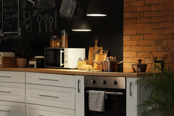 Interior of stylish kitchen with stove, utensils and glowing lamps at night