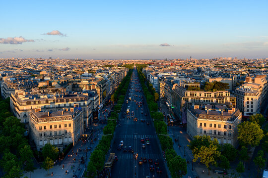 Aerial perspective of the Champs Elysees stretching through central Paris, flanked by classic Haussmann buildings and lush green trees under clear blue skies at sunset.