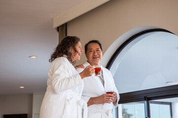 Senior couple standing in spa lounge wearing bathrobes by window holding glass mugs of tea