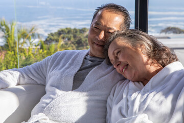 Senior couple relaxing in lounge on sofa wearing bathrobes by glass window overlooking hills