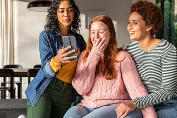 Diverse female friends gathering around smartphone sitting on sofa in living room near table