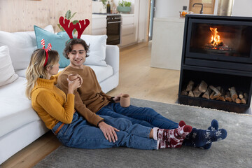 Naklejka premium Couple sitting on gray rug in living room beside fireplace holding mugs wearing antler headbands