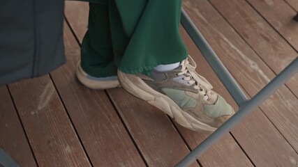 green pants sneaker under table closeup showing casual trainer and metal chair leg, cropped view of foot tapping and subtle toe touch between seated people on wooden deck, relaxed playful mood, soft