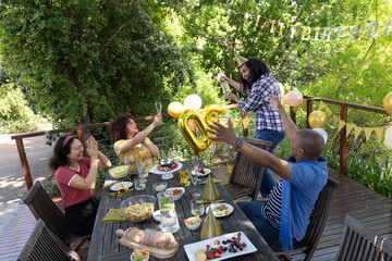 Diverse friends celebrating birthday on backyard deck around table with cake, champagne