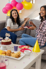 Diverse mom and teenage daughter sitting on sofa celebrating with red birthday cake and balloons