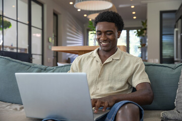 African american man working remotely on laptop on teal sofa in living room, copy space