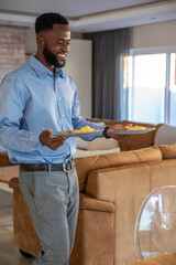 African american man carrying two plates of scrambled eggs, toast in living room, copy space