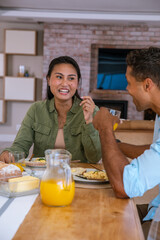 Diverse couple sitting at home sharing plates of scrambled eggs on table with juice jug