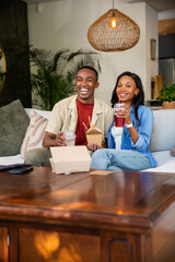 African American couple sitting on pale sofa in living room, holding take-out and sipping drinks
