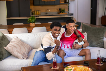 Diverse male friends celebrating and watching match on sofa in living room with pizza, soccer ball