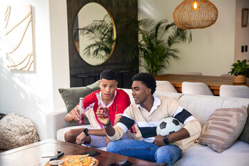Diverse male friends in red jersey, striped polo sitting on sofa at home with soccer ball