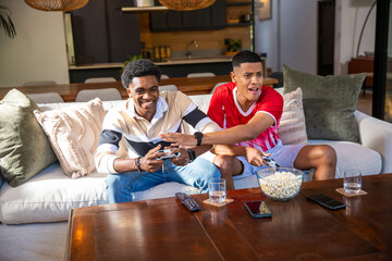 Diverse male friends holding game controllers and grabbing popcorn from coffee table in living room