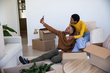 Diverse couple taking selfie with smartphone in living room among boxes while holding coffee cups