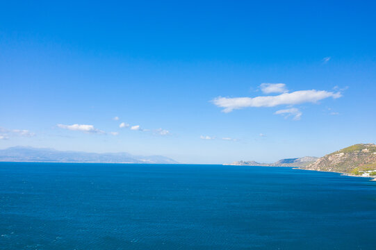 Wide view of a deep blue sea stretching toward distant mountains, with sunlit hills and scattered clouds beneath a vibrant, cloudless sky. The scene feels open, serene, and full of natural clarity.