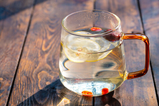 A clear glass cup holds warm water with lemon slices and goji berries. Sunlight shines on the drink, highlighting the wooden table in Chengdu, Sichuan, China.