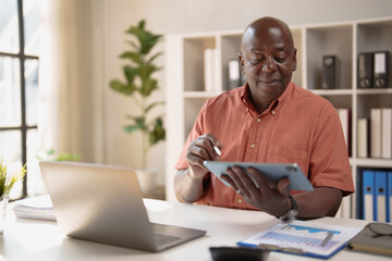 Fototapeta premium Mature African American businessman analyzing data on a digital tablet while working remotely from a modern home office, symbolizing modern work, technology, and success