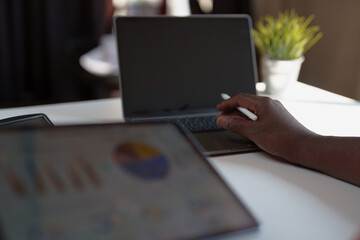 Person's hand holding a stylus, working on a laptop with a blank screen and a tablet displaying business charts in the foreground, representing digital work and data analysis