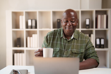 Smiling senior African American man holding a coffee mug, engaged in remote work using a laptop at his home office, enjoying connectivity and a comfortable workspace