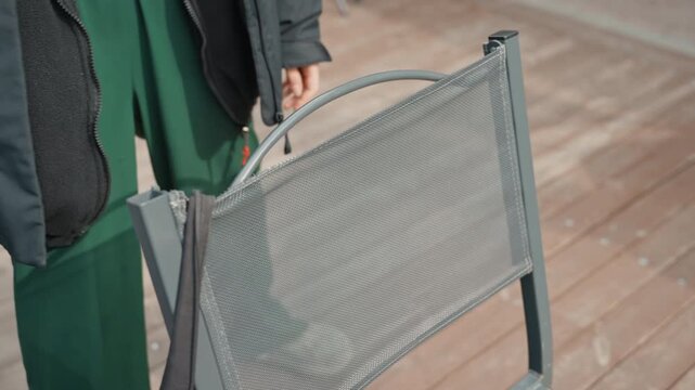 person adjusting metal patio chair, green trousers and dark coat, closeup on mesh backrest and steel frame, wooden deck planks underfoot, gentle hand movement, candid terrace atmosphere, soft natural