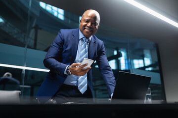 Smiling black mature businessman holding smartphone while standing at office desk, combining modern technology with professional leadership in a contemporary corporate environment
