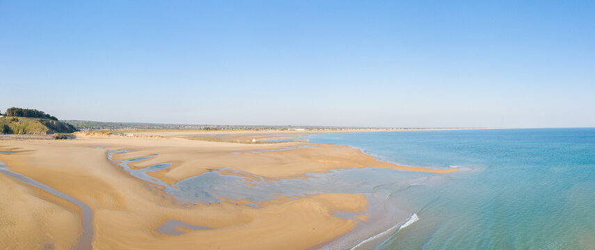 Vast sandy beach at Plage de la Potiniere stretches along calm turquoise water, featuring gentle tidal pools and a distant headland under clear blue sky. The scene is bright, peaceful, and open