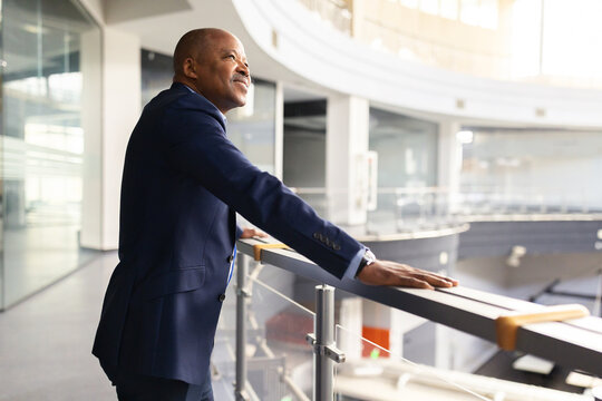 Black mature businessman leaning on office balcony railing, looking ahead with confidence and vision, executive lifestyle in modern corporate architecture