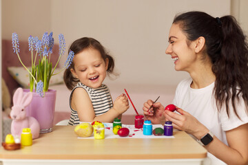 Happy mother and cute little child girl are preparing to celebrate Easter at home. Mom and daughter painting eggs before Easter together.