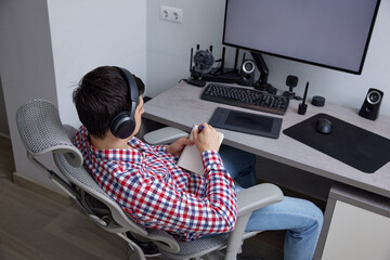 Man wearing headphones sitting at an ergonomic desk, writing notes in a notebook