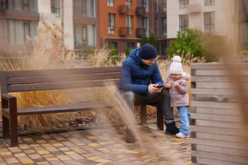 Father and daughter interacting with mobile phones while sitting on a bench in an urban park