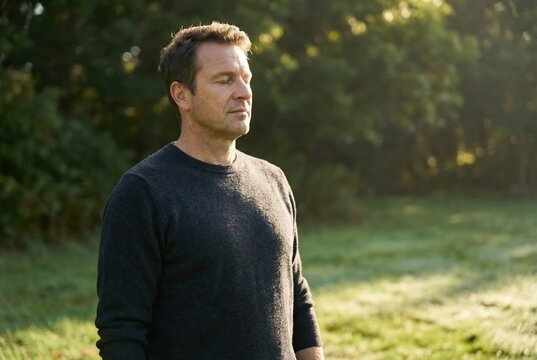A man is standing still in a green field, enjoying the morning light. He takes a moment for himself surrounded by nature and trees. The scene shows a peaceful morning setting