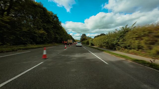 Car driving through road construction zone with traffic cones and repair equipment POV. Automobile moving along highway under maintenance surrounded by trees and cloudy sky first person view. SUV