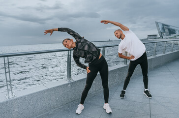 A man and woman include dynamic stretching in their routine by a riverside walkway, embracing active lifestyles.