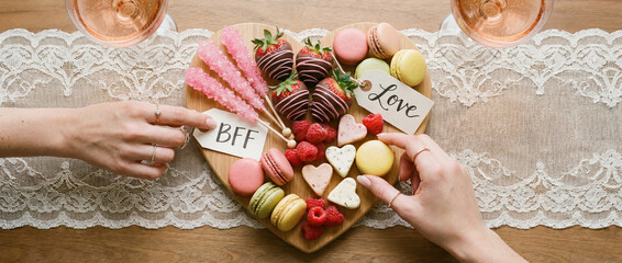 Naklejka premium Two women hands with rings share heart shaped dessert board with macarons, strawberries, raspberries, sugar rods, for Galentines Day celebration.