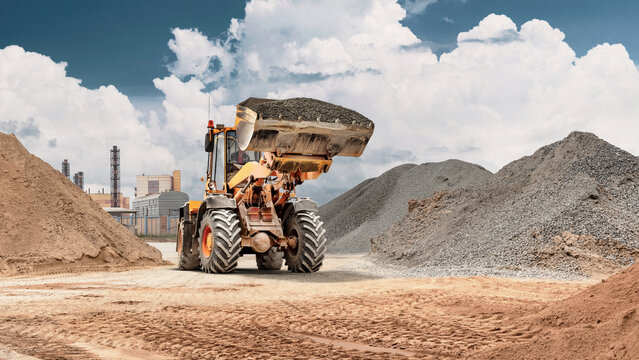 A wheel loader moves sand in a storage bucket at a construction site with piles of material and an industrial backdrop. Heavy machinery is in operation