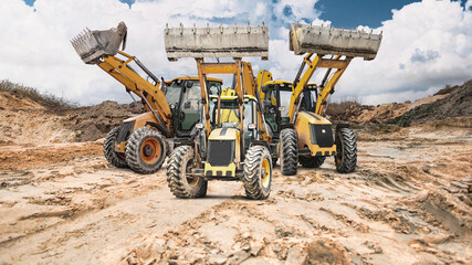 Three loaders with raised buckets are positioned at a construction site with earthen embankments or a quarry. The sky is partly cloudy, indicating a change in the weather