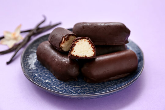 Plate with tasty chocolate glazed curd snacks with vanilla flavor on lilac background