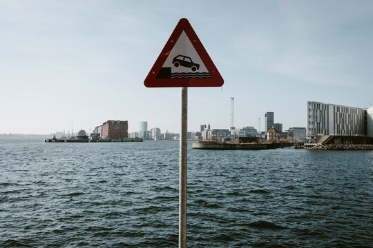 Waterfront harbor coast sign with warning triangle signals danger for vehicle access with sea cityscape backdrop supporting safe coastal mobility