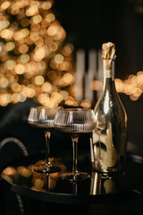 A closed bottle of champagne or sparkling wine stands next to two glass glasses against the background of a Christmas tree decorated with toys and garlands