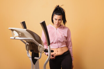 A young woman in sportswear stands by an elliptical trainer and gently shows loose stomach skin against a solid yellow background. This image represents fitness journey, weight loss results, 