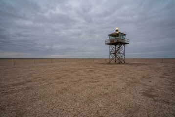 Fototapeta premium A watchtower is set in a barren area with cracked earth and low vegetation. The sky is full of clouds, and the watchtower looks over a distant body of water