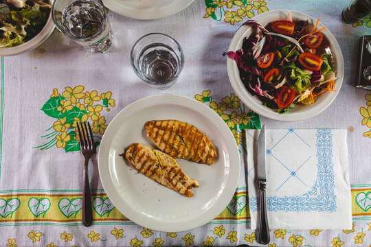 piatto con fette di petto di pollo e scodella con insalata mista e pomodori, su un tavolo, con posate e bicchieri riempiti con dell'acqua, piatto tradizionale estivo italiano leggero e salutare