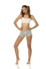 Fototapeta premium Studio shot of young woman in white top and grey shorts smiling with hands on hips on white background, full body