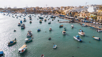 Aerial drone view of Marsaxlokk harbor in Malta. Traditional colorful Luzzu fishing boats in turquoise water. Ideal for tourism marketing, travel guides, and Mediterranean lifestyle blogs.
