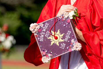 A high school or college graduate holds her decorated cap reading "now's when my life begins" &mdash; a joyful, personal image of commencement, achievement, and life's next chapter.